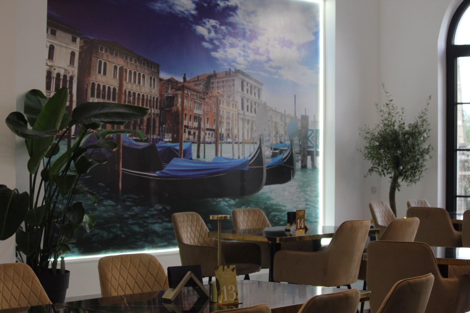 Un mural pictat manual cu gondole venețiene ancorate pe un canal, văzut printre un măslin și o masă de restaurant din interiorul Giovanni's